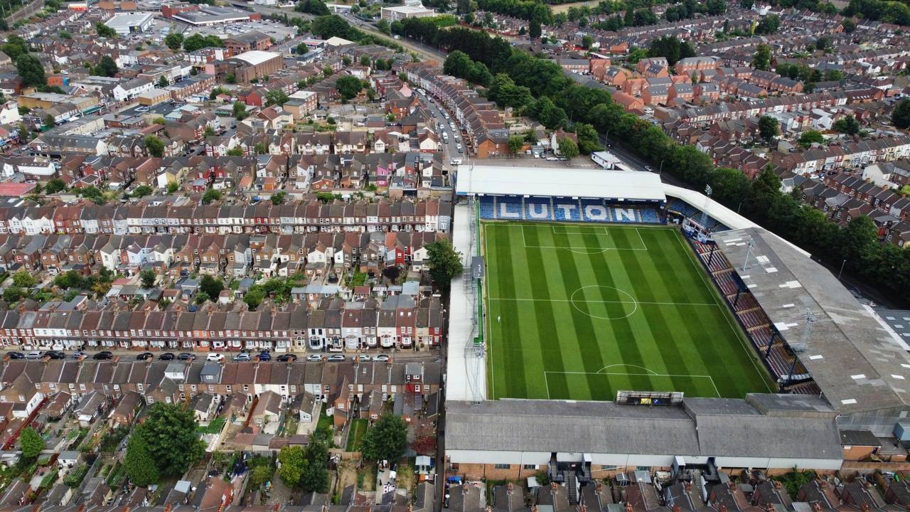 Vista aérea de estádio de futebol com "LUTON" nas arquibancadas, cercado por casas urbanas.