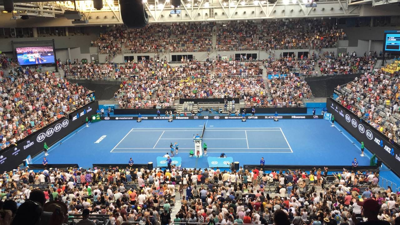 Quadra de tênis azul com jogadores, árbitro, telões e público lotado em estádio indoor. Logos: Kia, Mastercard, Hisense.