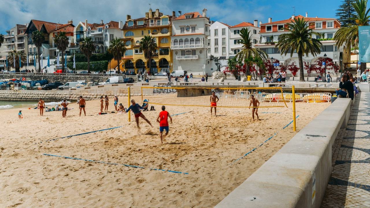 Homens jogando vôlei de praia na areia, com rede amarela e edifícios à beira-mar.