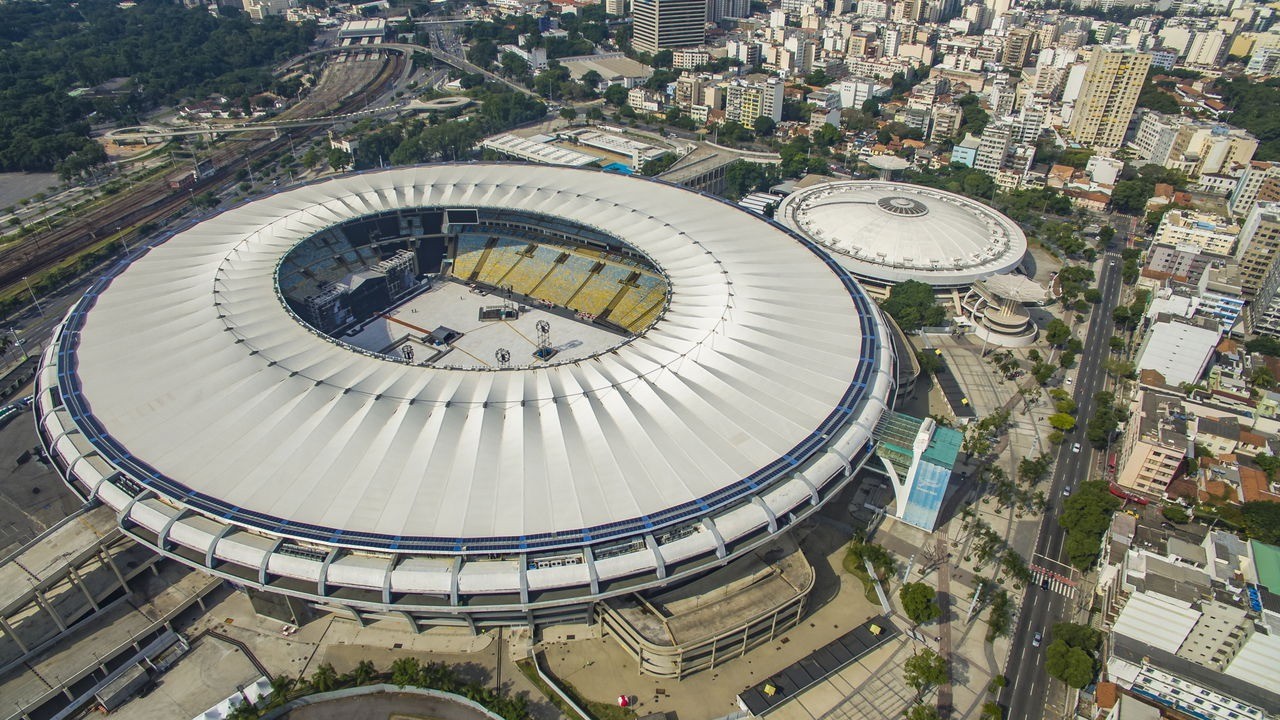 Vista aérea de estádio de futebol circular com teto branco, assentos internos e cidade ao redor.