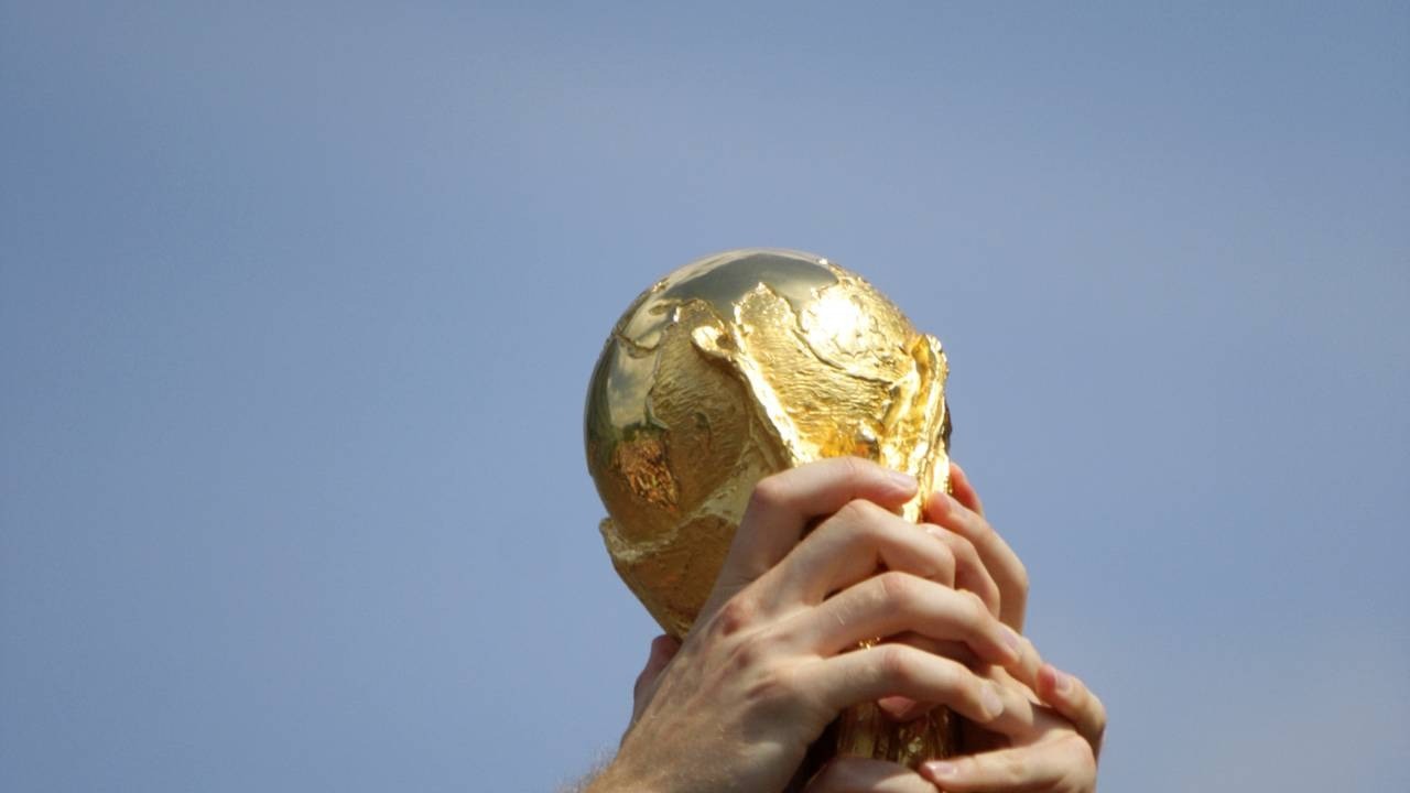 Taça dourada da Copa do Mundo sendo segurada por mãos contra um céu azul claro.