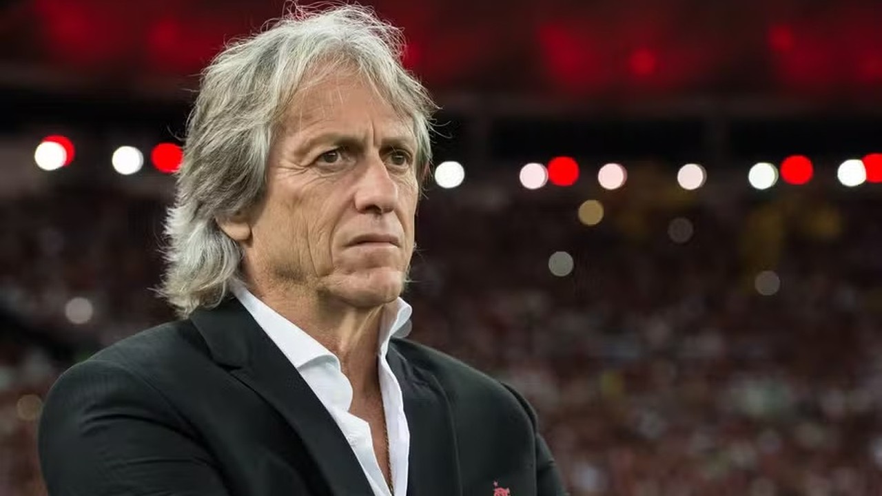 Jorge Jesus, técnico português, em close-up, vestindo blazer preto com fundo desfocado de estádio.