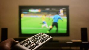 Mão segura controle remoto; TV desfocada exibe partida de futebol com jogadores e bola.