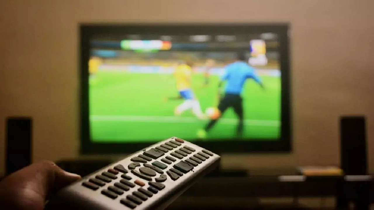 Mão segura controle remoto; TV desfocada exibe partida de futebol com jogadores e bola.