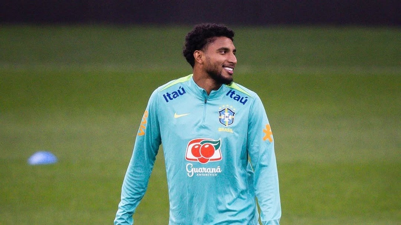 Jogador Éderson com camisa de treino da Seleção Brasileira, sorrindo em campo de futebol.